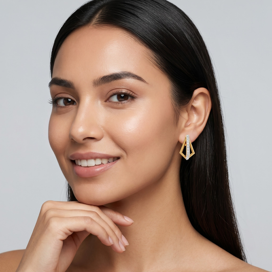 A close-up of a woman with long dark hair wearing a gold geometric dangle earring, set against a solid light gray background.