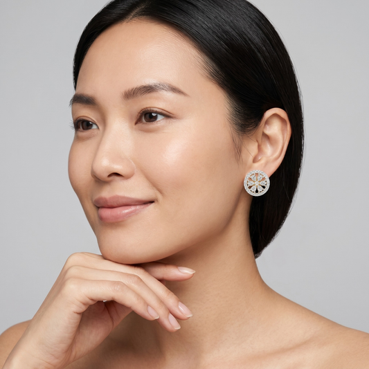 A close-up portrait of a woman with dark hair pulled back, wearing a single round stud earring with a silver-toned floral or sunburst design and clear stones, against a light gray studio background.