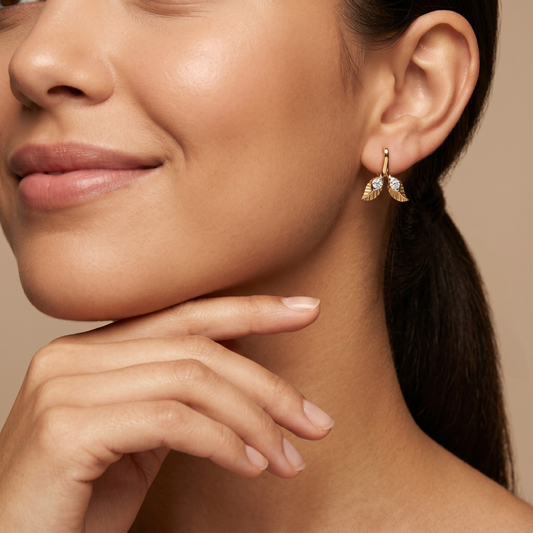A close-up lifestyle shot of a woman wearing a delicate gold leaf-shaped drop earring with small clear stones, set against a warm beige background.