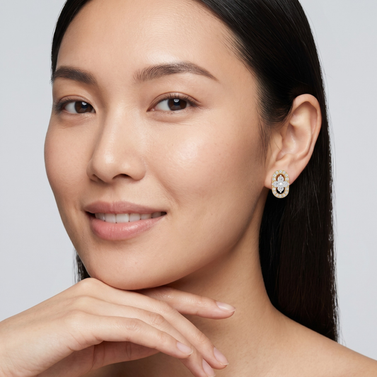 A close-up portrait of a woman wearing a small gold and white gemstone stud earring, with her hand resting near her chin against a light gray background.