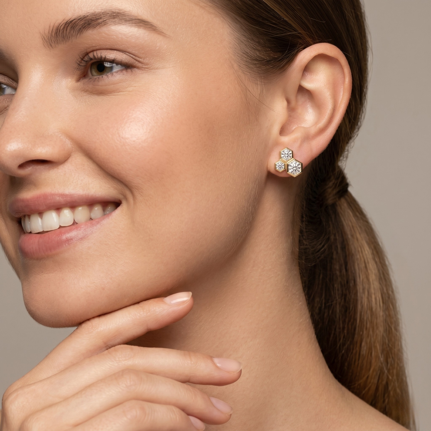 A close-up of a woman wearing a small four-petal flower-shaped stud earring with clear gemstones in a silver-toned setting, against a soft warm gray background.