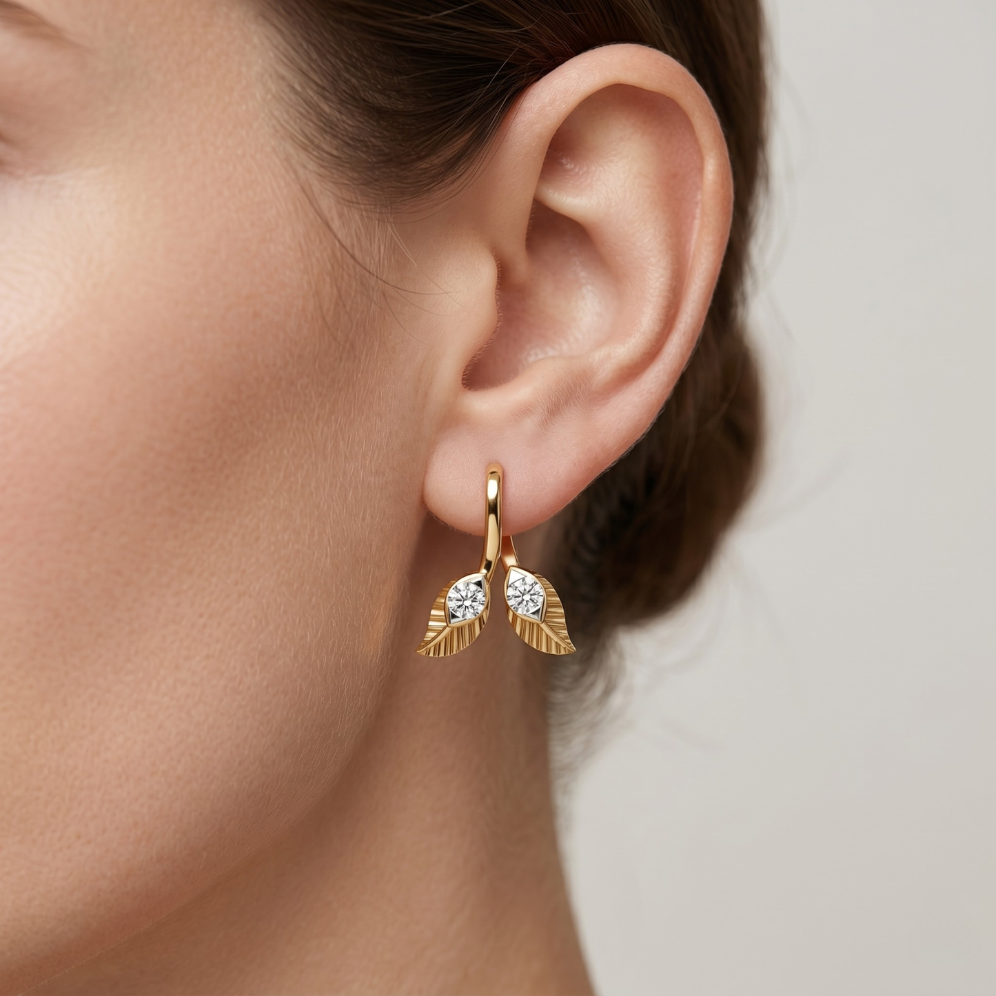 A close-up of a model's ear wearing a gold huggie hoop earring with two leaf-shaped drops, each set with a clear round gemstone.
