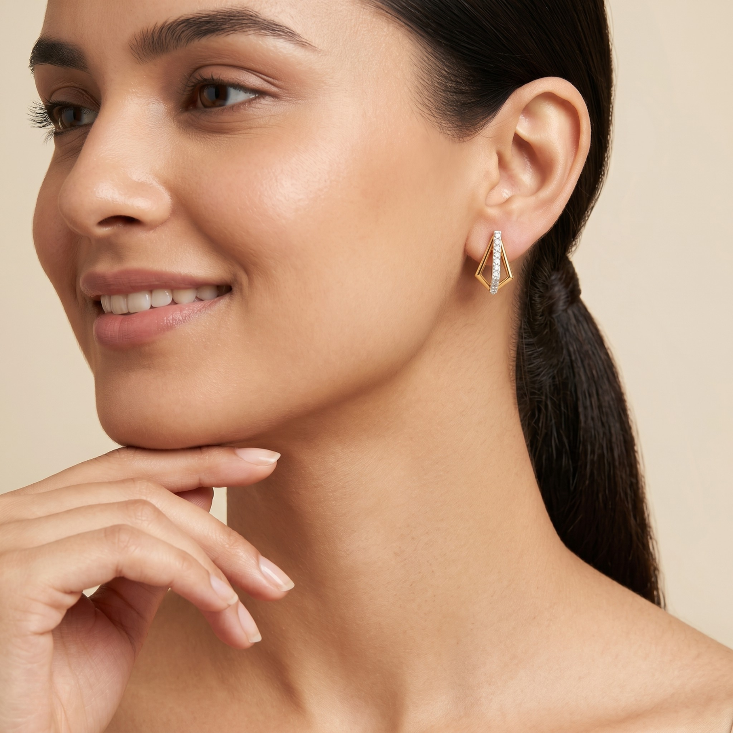 A close-up of a woman wearing a small gold geometric dangle earring with clear stones, set against a warm beige background.