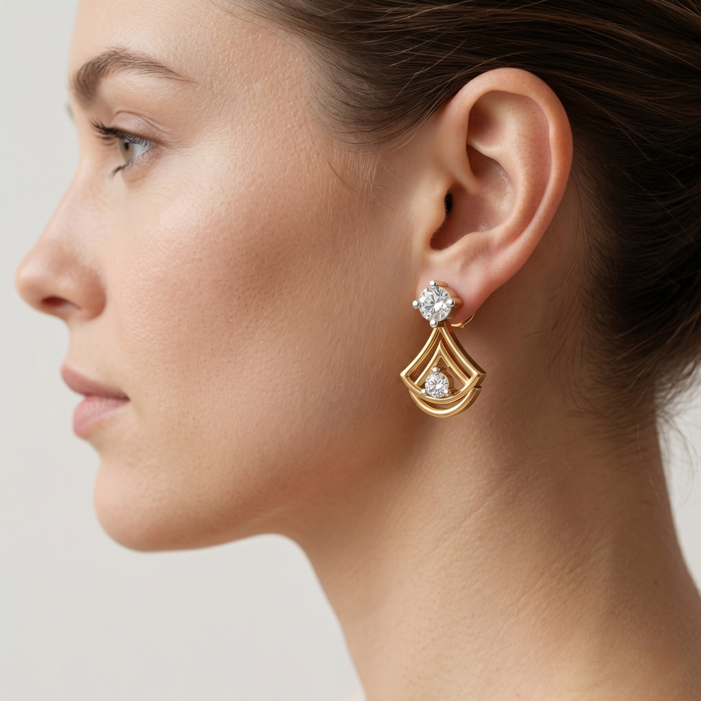 A close-up side profile of a woman wearing a gold drop earring with clear gemstones, set against a soft light gray background.