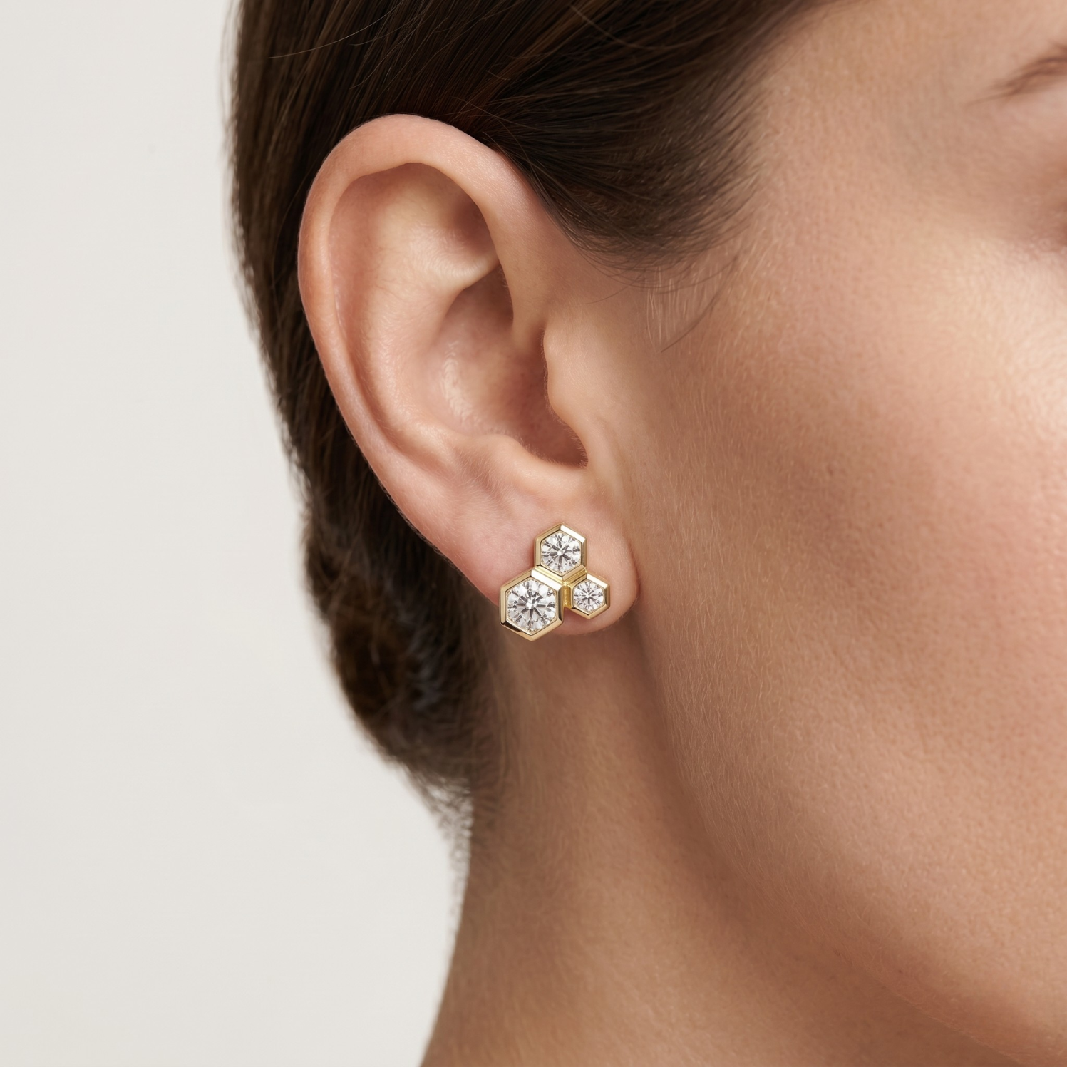 A close-up of a model's ear wearing a small gold stud earring with three clear faceted gemstones clustered together.