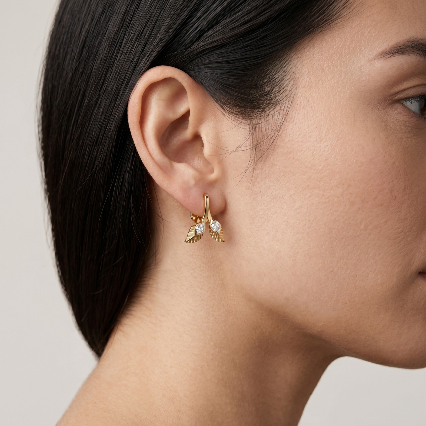 A close-up side profile of a woman wearing a delicate gold huggie hoop earring with a small gold leaf and clear crystal charm, set against a soft light gray background.