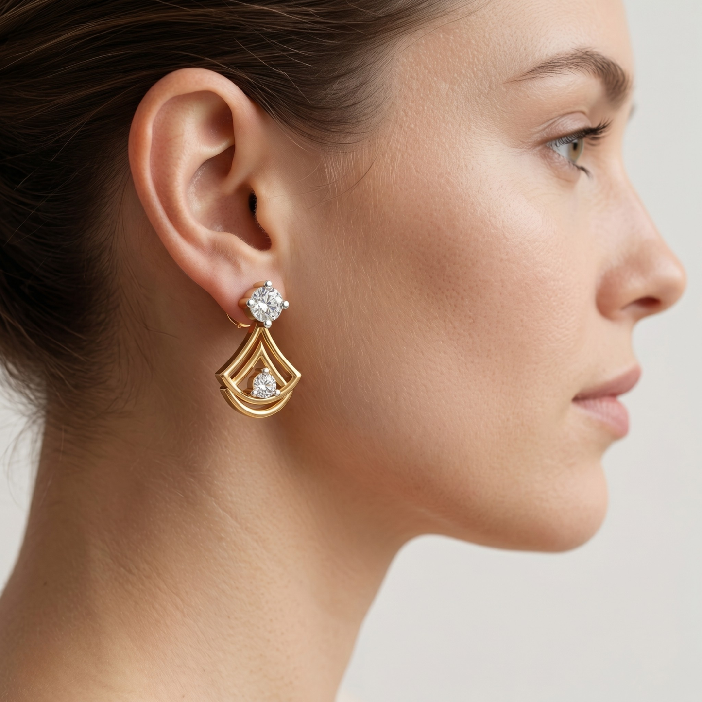 A close-up side profile of a woman wearing a gold drop earring with clear gemstones, set against a soft light gray background.
