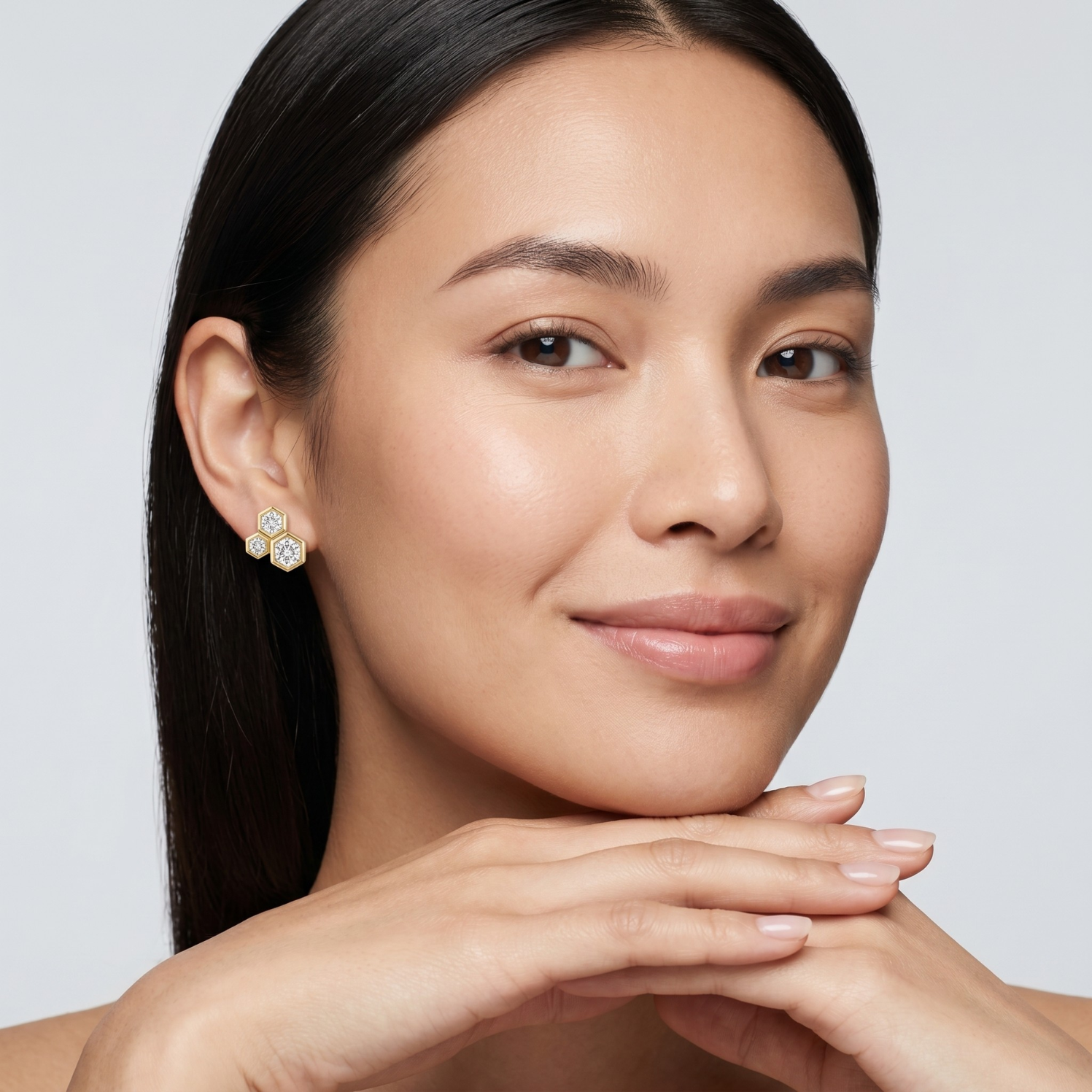 A close-up portrait of a woman with dark hair wearing a small gold and white floral stud earring, with her hands resting near her chin against a light gray background.