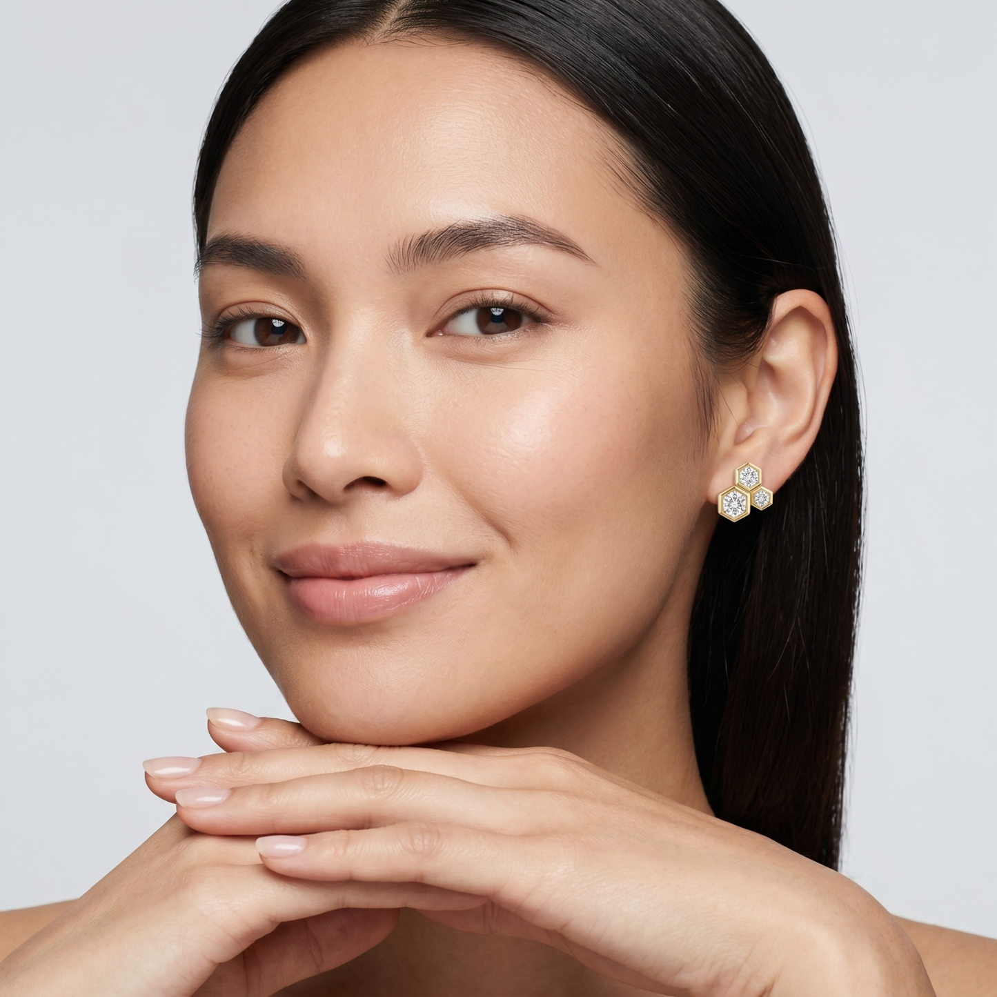 A close-up portrait of a woman wearing a small gold flower-shaped stud earring with clear gemstones, set against a clean light gray background.