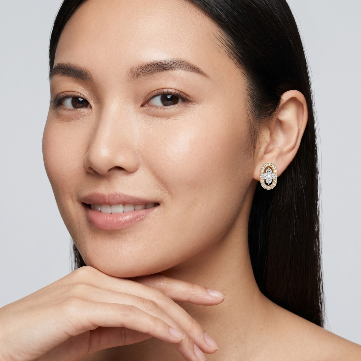 A close-up portrait of a woman wearing a small gold and white gemstone stud earring, with her hand resting near her chin against a light gray background.